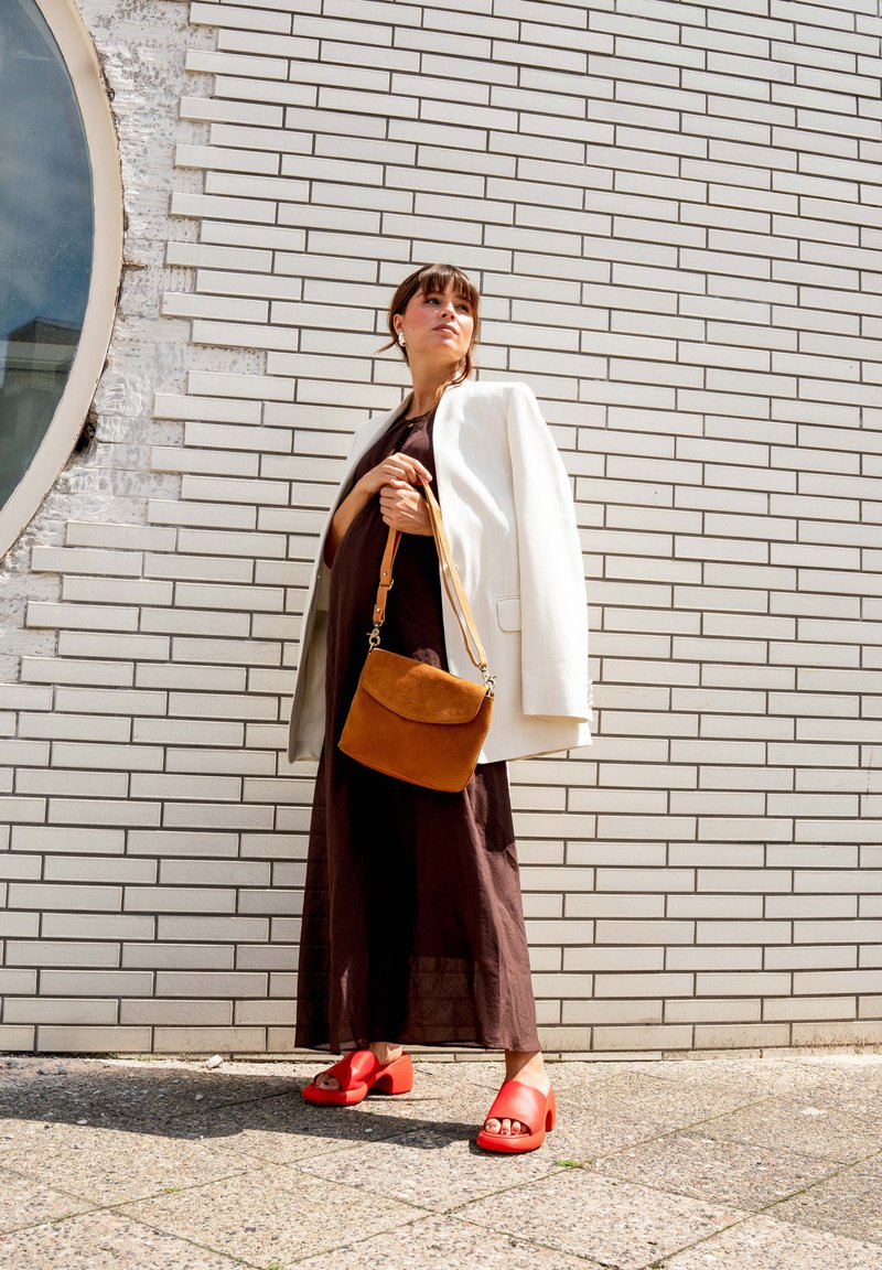 Frau in braunem Kleid und weißem Blazer steht vor einer Backsteinmauer, hält eine beigefarbene Handtasche und trägt rote Plateau-Sandalen.
