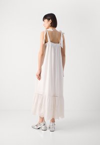 Vestido blanco sin mangas con lazos en los hombros, una parte trasera fruncida y un dobladillo con encaje. Combinado con zapatos de tacón bloque plateados.
