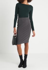 Haut vert foncé à manches longues avec un large décolleté, assorti à une jupe crayon grise. Le mannequin tient un sac à main noir et porte des bottes noires montant aux genoux.