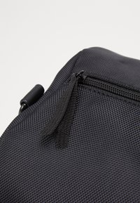 Bolso negro texturizado con cierre de cremallera, que presenta una pequeña pestaña. El material parece duradero y estructurado, con un patrón geométrico.