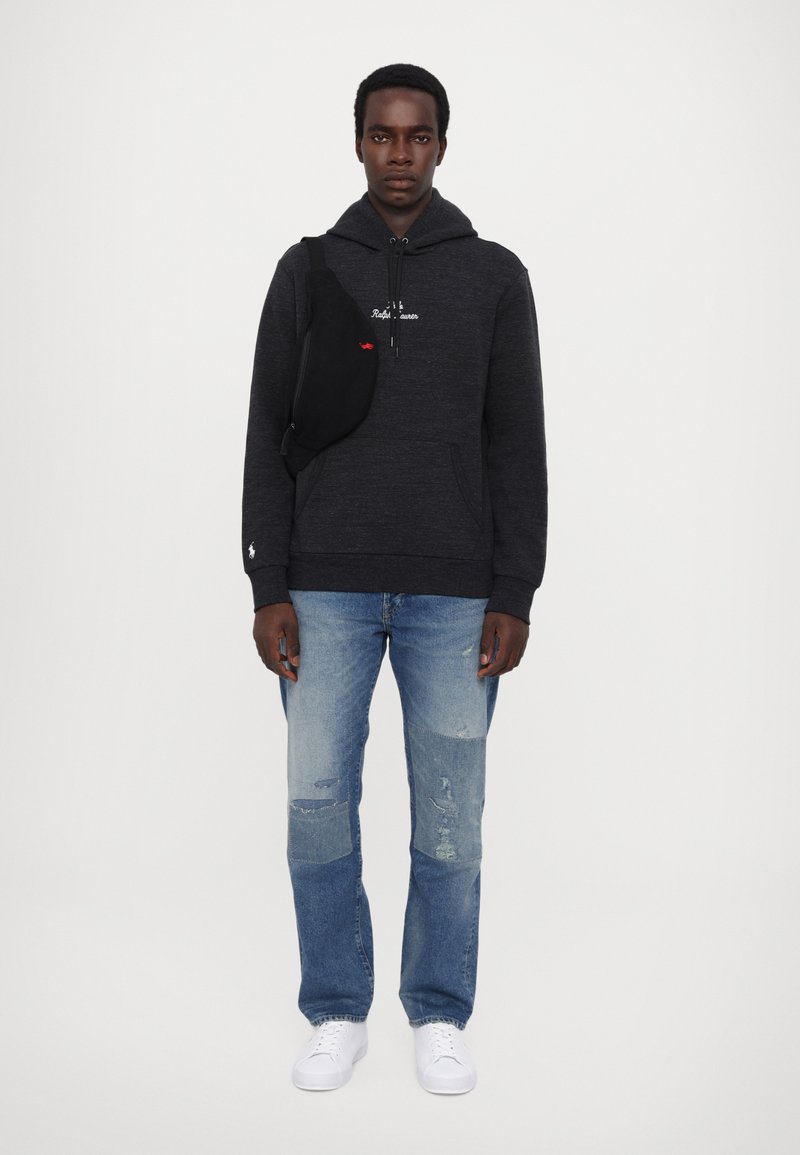 Schwarzer Hoodie mit Fronttasche und weißem Logo, kombiniert mit hellblauen, ausgewaschenen Jeans und weißen Sneakers. Verstellbare schwarze Umhängetasche.