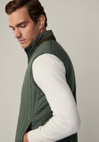 Gestepptes grünes Vest mit hohem Kragen, das einen braunen Akzent an der Schulter hat. Trägt man über einem weißen Langarmshirt.