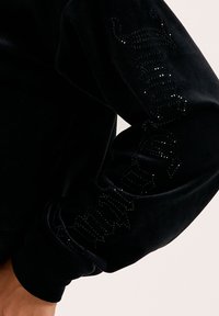 Schwarzes Samtoberteil mit strukturiertem Design und Strassakzenten entlang des Ärmels, das eine weiche Oberfläche und eine enganliegende Passform präsentiert.