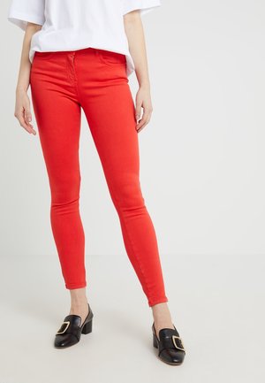 Person trägt leuchtend rote Skinny-Hose, weißes Oberteil und schwarze Pumps mit großen goldenen Schnallen, steht auf einem schlichten hellen Hintergrund.
