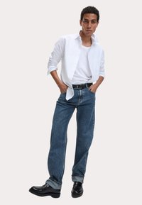 Camisa blanca de botones, camiseta blanca debajo, vaqueros de mezclilla azules, cinturón negro y zapatos negros. Los vaqueros están ligeramente remangados en los puños.