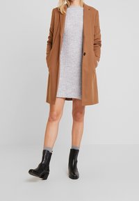 Cappotto in misto lana marrone, vestito grigio a coste, calzini grigi, stivaletti neri alla caviglia con tacco a blocco, in piedi su uno sfondo neutro.