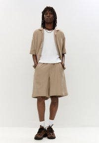 Chemise boutonnée à manches courtes en tricot beige sur un T-shirt blanc, associée à un short beige assorti et des accents marron sur des baskets.