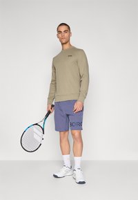 Light olive long-sleeve sweatshirt, blue shorts with "BORG" print, white sports shoes, holding a tennis racquet, neutral background.