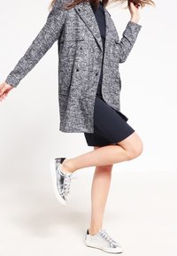 Cappotto grigio testurizzato doppiopetto su un vestito blu navy, abbinato a scarpe da ginnastica argentate con suola bianca.