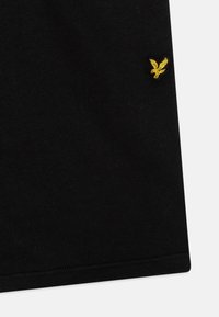 T-shirt en coton noir avec un petit logo d'aigle brodé en jaune. Texture lisse, manches courtes et coupe standard.