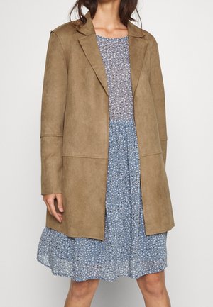 Mujer con un blazer de ante marrón claro sobre un vestido azul con flores hasta la rodilla, de pie frente a un fondo blanco liso.