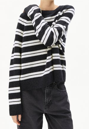 Personne portant un pull rayé horizontal noir et blanc et un jean noir, avec un bras plié et la main reposant près de l'épaule.