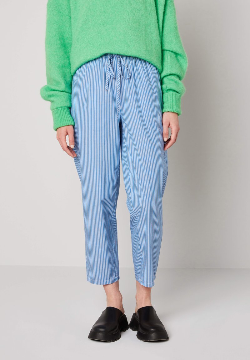 Inimene, kes kannab helesinist ja valget triibulist püksipaari, millel on nöörvöökoht, eredat rohelise värvi džemprit ning musti libisevaid kingi.