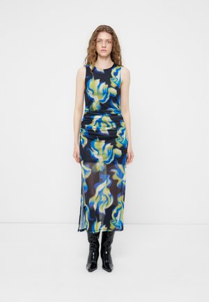 Femme aux cheveux ondulés portant une robe longue sans manches à motifs abstraits bleu, jaune et noir et des bottines noires, debout contre un mur uni.