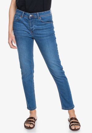 Personne portant un jean skinny bleu taille moyenne et des sandales à enfiler imprimé léopard, debout sur un fond blanc.