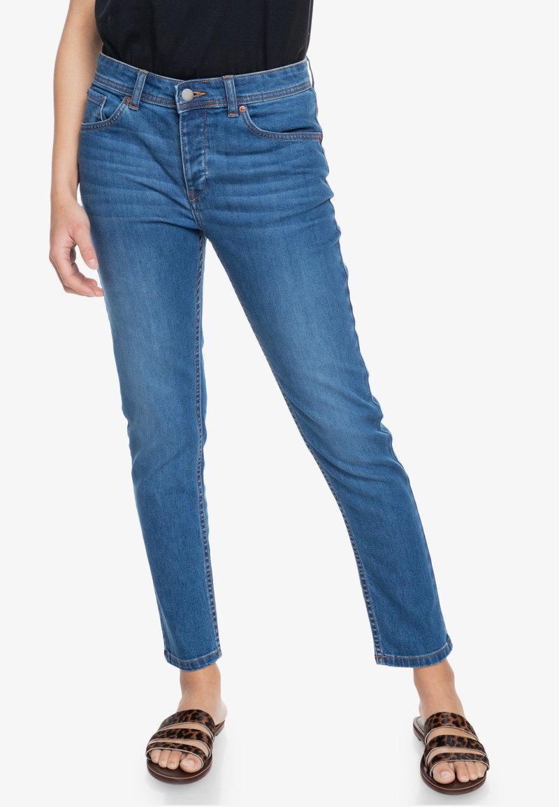 Personne portant un jean skinny bleu taille moyenne et des sandales à enfiler imprimé léopard, debout sur un fond blanc.