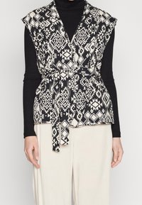 Svart och vit mönstrad väst med inslag av omslag, korta ärmar och knytbälte. Bärs över en svart långärmad tröja och krämfärgade byxor.