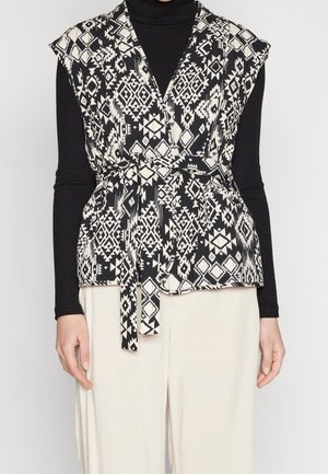 Schwarze und weiße gemusterte Weste mit Wickeldesign, kurzen Ärmeln und einem Bindegürtel. Getragen über einem schwarzen Langarmoberteil und cremefarbenen Hosen.