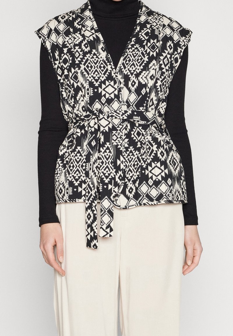 Svart och vit mönstrad väst med inslag av omslag, korta ärmar och knytbälte. Bärs över en svart långärmad tröja och krämfärgade byxor.