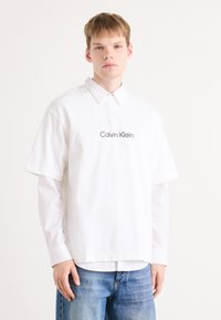 Balta didelio dydžio trumpomis rankovėmis marškiniai su taškuota apykakle, papuošti siuvinėtu "Calvin Klein" logotipu. Derinama su mėlynomis džinsinėmis kelnėmis.