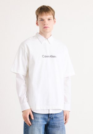 White oversize short-sleeve shirt with a point collar, featuring embroidered "Calvin Klein" logo. Paired with blue denim jeans.