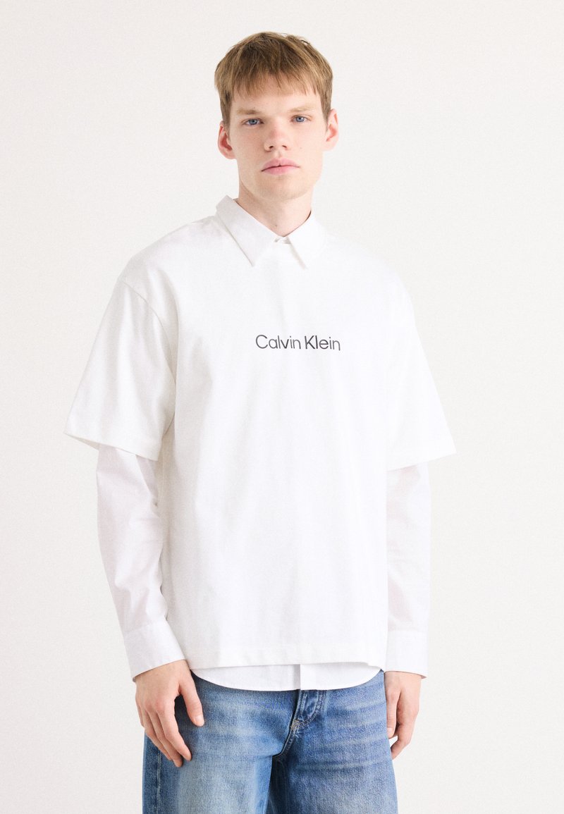 Balta didelio dydžio trumpomis rankovėmis marškiniai su taškuota apykakle, papuošti siuvinėtu "Calvin Klein" logotipu. Derinama su mėlynomis džinsinėmis kelnėmis.