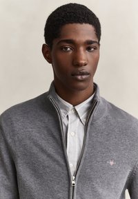 Joven con cabello corto y rizado que lleva una camisa blanca abotonada bajo un jersey gris con cremallera, mirando hacia adelante sobre un fondo liso.