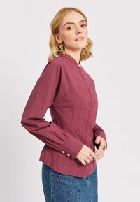 Donna che indossa un blouse aderente color bordeaux con dettagli in bottone dorato e orecchini d'oro statement, abbinata a una gonna di jeans blu.