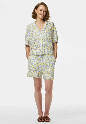 Donna sorridente, indossa una camicia gialla a maniche corte con bottoni e motivo floreale, pantaloncini abbinati con le mani nelle tasche, e sandali marroni.