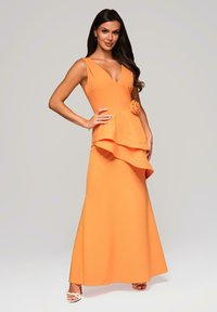 Vrouw staat vol vertrouwen in een mouwloze oranje jurk met een V-hals en asymmetrische ruches rond de taille, met witte hoge hakken aan.
