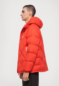 Red puffer jacket with a diamond quilt pattern, hood, front zipper and snap closure. Features elastic cuffs and a side pocket.