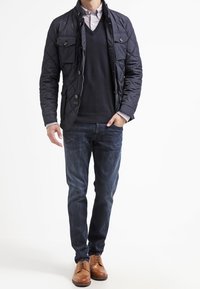 Marineblaue Steppjacke mit zwei Vordertaschen, darüber ein dunkler Pullover und ein gestreiftes Hemd. Kombiniert mit blauer Jeans und braunen Lederschuhen.