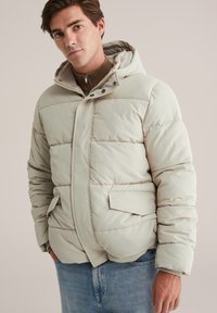 Chaqueta acolchada beige con capucha, con un patrón acolchado, botones a presión y bolsillos laterales. Textura suave y lisa con un corte relajado.