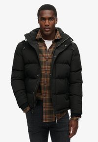 Czarna kurtka puchowa z kapturem, z zapięciem na zamek, elastycznymi mankietami i detalami na ramionach, noszona na koszuli w kratę z guzikiem.