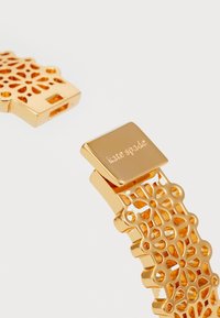 Gullfarget metallarmbånd med et utskåret blomsterdesign. Har en glatt, polert lås gravert med "kate spade." Teksturert og intrikat design.