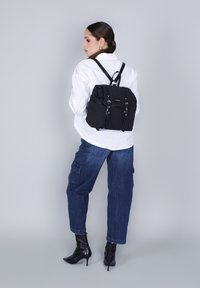 Mochila negra de nailon con textura suave, dos bolsillos frontales, correas ajustables y un diseño moderno, llevada en la espalda de una figura.