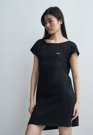 Γυναίκα με μακριά μαύρα μαλλιά που φοράει μαύρο φόρεμα με κοντά μανίκια και μικρό χρυσό λογότυπο «EA7» στο στήθος, στέκεται μπροστά σε απλό γκρι φόντο.