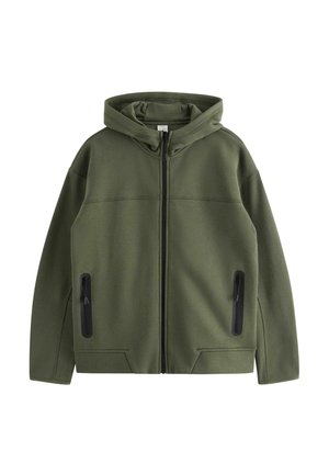 Boy standing with hands in olive green zip-up hoodie and matching joggers, navy blue shirt, and navy sneakers with white soles.
