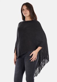 Poncho lavorato a maglia nero con una texture a coste, caratterizzato da un orlo frangiato e lati aperti. Il design include piccole aperture per la ventilazione.