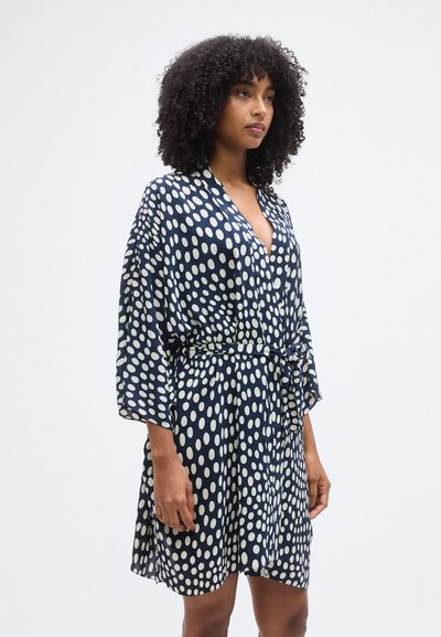 Μπλε ρουά φόρεμα με V-λαιμόκοψη, φαρδιά μανίκια και λευκές πουά βούλες. Έχει ζώνη στη μέση και ελαφρύ, αιωρούμενο ύφασμα.