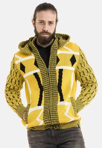 Gelbe Strick-Hoodie mit einem strukturierten, Kabelstrickmuster an den Ärmeln, das schwarze und weiße geometrische Designs am Körper enthält.