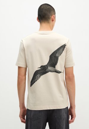 Mann trägt cremefarbenes T-Shirt mit großem schwarzem und grauem Vogelmotiv, das auf dem Rücken die Flügel ausbreitet, kombiniert mit dunklen Jeans.