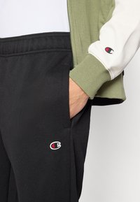 Pantalon de survêtement noir avec une texture subtile, doté de poches latérales et d'un petit logo brodé. Veste vert olive avec un détail blanc sur la manche.