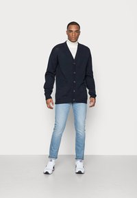 Marineblaue Strickjacke mit V-Ausschnitt und fünf Knöpfen, kombiniert mit hellblauen Jeans. Die Strickjacke hat gerippte Bündchen und zwei Fronttaschen.