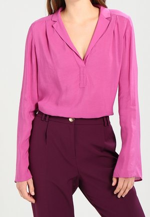 Vrouw draagt een roze blouse met lange mouwen en V-hals en een donkerpaarse hoog getailleerde broek met een gouden knoop.