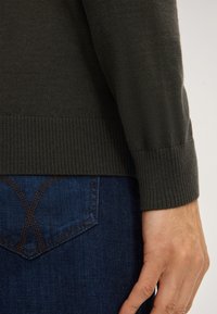 Maglione a maglia verde scuro con polsini a costine, indossato sopra jeans blu scuro caratterizzati da un design di tasche triangolari distintive e cuciture a contrasto.