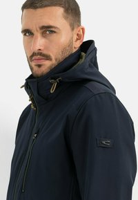 Parka bleu marine avec fermeture éclair, capuche ajustable et cordons décoratifs. Inclut un badge sur la manche gauche.