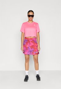 Ροζ κομμένο αθλητικό μπλουζάκι με κοντά μανίκια και λογότυπο, σε συνδυασμό με πολύχρωμη φούστα με λουλουδάτο μοτίβο σε αποχρώσεις του ροζ και του μωβ.