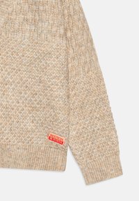 Beige strikkegenser med diamantmønster, ribbestrikkede mansjetter og et rødt merke med "SCOTCH & SODA" på kanten.