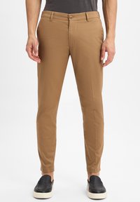 Man in een slim-fit lichtbruine broek en zwarte instapschoenen met witte zolen, staand tegen een eenvoudige lichte achtergrond.